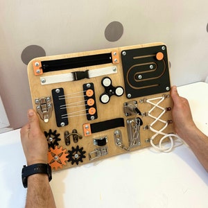 May include: A wooden busy board with various activities for children, including a zipper, a buckle, a latch, a chain, a spinner, a maze, and a lacing activity. The board is painted in natural wood tones with black and orange accents.