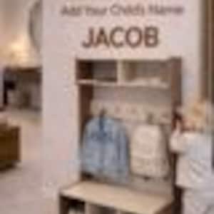 May include: A light brown wooden entryway storage unit featuring an upper shelf, a middle section with hooks for hanging items, and a bottom bench with three open cubbies for shoe storage. The text "Free Personalization Add Your Child's Name JACOB" is visible.