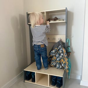 May include: A tall, gray and light wood entryway storage unit featuring an upper shelf, a middle section with three metal coat hooks, and a lower bench with three open cubbies. A patterned jacket hangs from a hook, and a teal umbrella is attached.