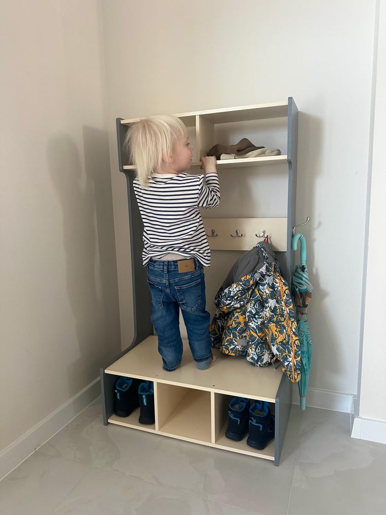 May include: A tall, gray and light wood entryway storage unit featuring an upper shelf, a middle section with three metal coat hooks, and a lower bench with three open cubbies. A patterned jacket hangs from a hook, and a teal umbrella is attached.