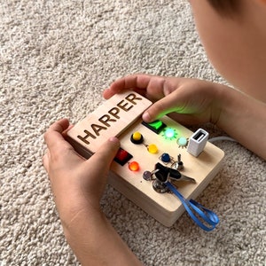 Peut inclure: Un tableau d'activités en bois avec le nom "HARPER" gravé sur le dessus. Le tableau propose une variété d'activités, notamment des boutons, des interrupteurs, un trou de serrure et un port USB. Le tableau est de couleur brun clair et a une finition en bois naturel.