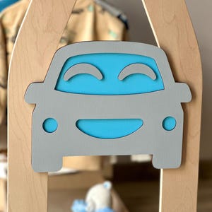 May include: A wooden children's playhouse with a gray car-shaped cutout on the front. The car has a blue interior and smiling face design. The playhouse is made of light-colored wood.