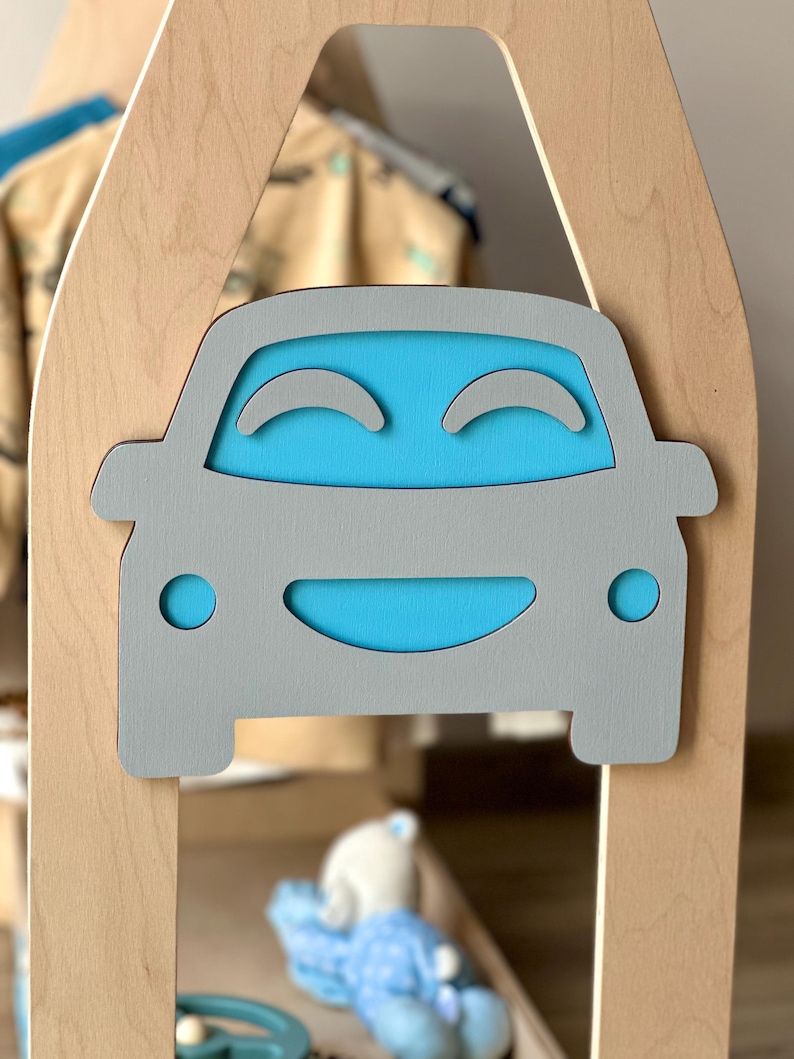 May include: A wooden children's playhouse with a gray car-shaped cutout on the front. The car has a blue interior and smiling face design. The playhouse is made of light-colored wood.