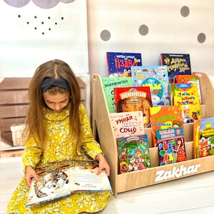 Puede incluir: Una estantería de madera con una etiqueta blanca que dice "Zakhar" y está llena de libros infantiles coloridos. Una niña está sentada en el suelo frente a la estantería, leyendo un libro.