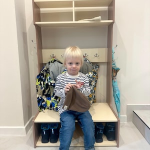May include: A light-colored wooden entryway hall tree featuring an upper storage shelf, a central row of metal coat hooks, and a lower bench with an open shoe storage compartment underneath. The unit is designed for organizing coats, hats, and footwear.