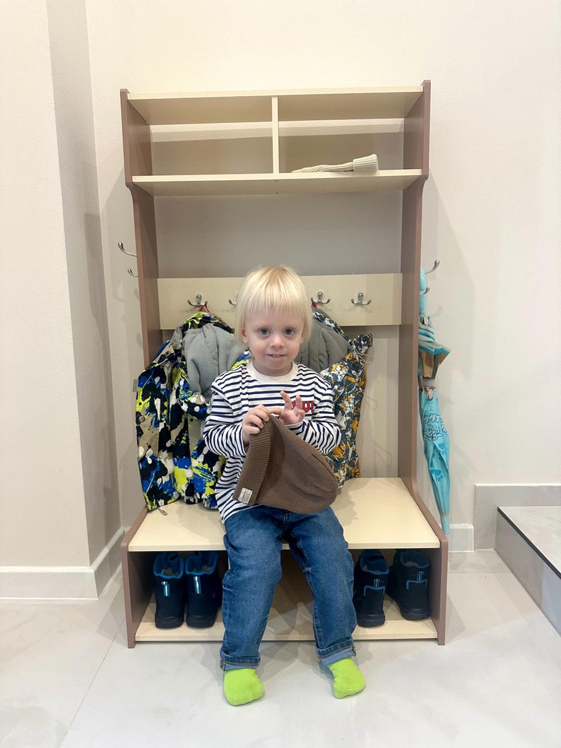 May include: A light-colored wooden entryway hall tree featuring an upper storage shelf, a central row of metal coat hooks, and a lower bench with an open shoe storage compartment underneath. The unit is designed for organizing coats, hats, and footwear.