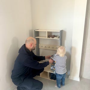 May include: A light-colored wooden entryway organizer featuring a top shelf, a middle section with five small metal hooks for hanging items, and a bottom storage area with two open cubbies, framed by two vertical grey side panels.