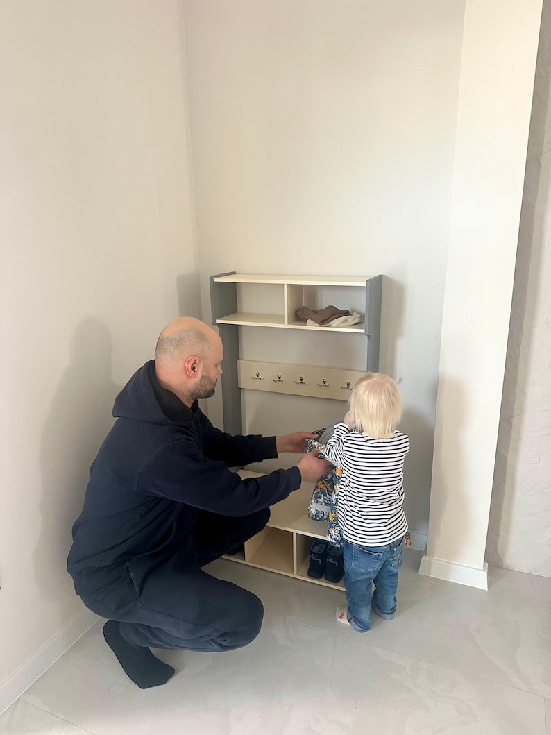 May include: A light-colored wooden entryway organizer featuring a top shelf, a middle section with five small metal hooks for hanging items, and a bottom storage area with two open cubbies, framed by two vertical grey side panels.