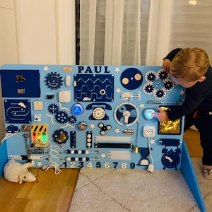 Può includere: Un pannello di attività in legno blu e bianco con varie texture e attività per bambini. Il pannello presenta un telefono, ingranaggi, un interruttore della luce, una campana, una serratura, una porta, uno slider e i numeri da 1 a 9. Il pannello è personalizzato con il nome "PAUL".