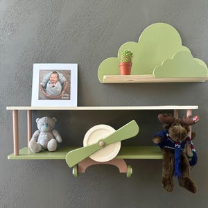 May include: A green aeroplane-shaped shelf with a teddy bear and a moose plush toy. The shelf is mounted on a wall with a green cloud-shaped shelf above it. The cloud shelf has a small cactus plant on it. A framed photo of a baby is on the wall below the aeroplane shelf.