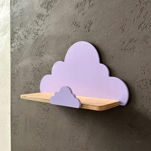 May include: A lavender cloud-shaped shelf with a smaller cloud detail on the front. The shelf is made of wood and is mounted on a wall.