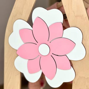 May include: A wooden decorative piece featuring a large, layered flower. The flower has pink petals with a white base, and a white center. The flower is attached to a light-colored wooden frame, creating a layered effect.