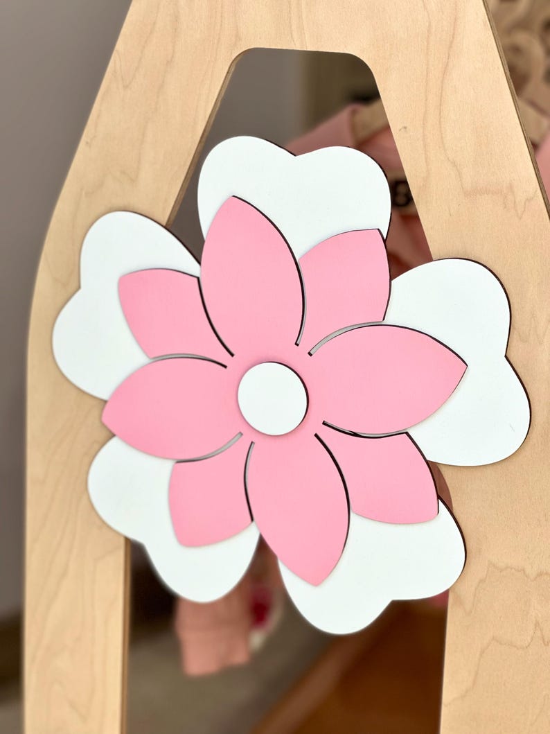 May include: A wooden decorative piece featuring a large, layered flower. The flower has pink petals with a white base, and a white center. The flower is attached to a light-colored wooden frame, creating a layered effect.