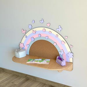 May include: A wooden rainbow-shaped shelf in pastel colors with a corkboard center. Decorative butterflies, flowers, and hearts surround the rainbow. A small gift box, a puzzle, and a lavender purse sit on the shelf.