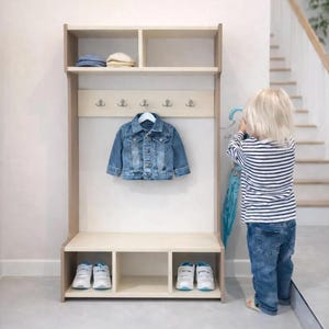 May include: A light-colored wooden entryway storage unit featuring an upper shelf with two compartments, a central panel with five silver hooks, and a lower bench with three open cubbies. A blue denim jacket hangs from one of the central hooks.