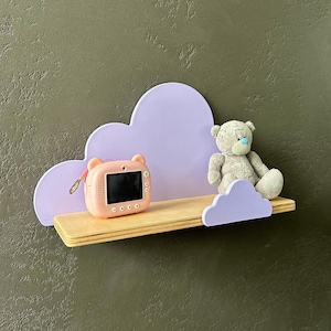 Pode incluir: Uma prateleira em forma de nuvem de cor lilás claro com uma base de madeira. Uma câmera de brinquedo rosa e um urso de pelúcia cinza estão sentados na prateleira.