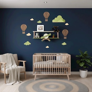 Pode incluir: Um berçário com uma parede azul marinho decorada com balões de ar quente de madeira, nuvens e uma prateleira com um ursinho de pelúcia e um avião. Um berço de madeira, uma poltrona e um tapete redondo completam o quarto. Uma planta em vaso está no canto.