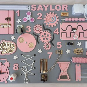 May include: A gray wooden sensory board with pink and silver elements. Features include a door, gears, a spinner, numbers, a clock, and the name "SAYLOR". Includes a variety of textures and interactive components for sensory exploration.