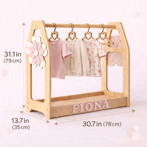 May include: A wooden children's clothing rack, 30.7 inches (78 cm) wide and 31.1 inches (79 cm) tall, with a flower accent and the name "FIONA" in pink letters. It holds several small garments on heart-shaped hangers.