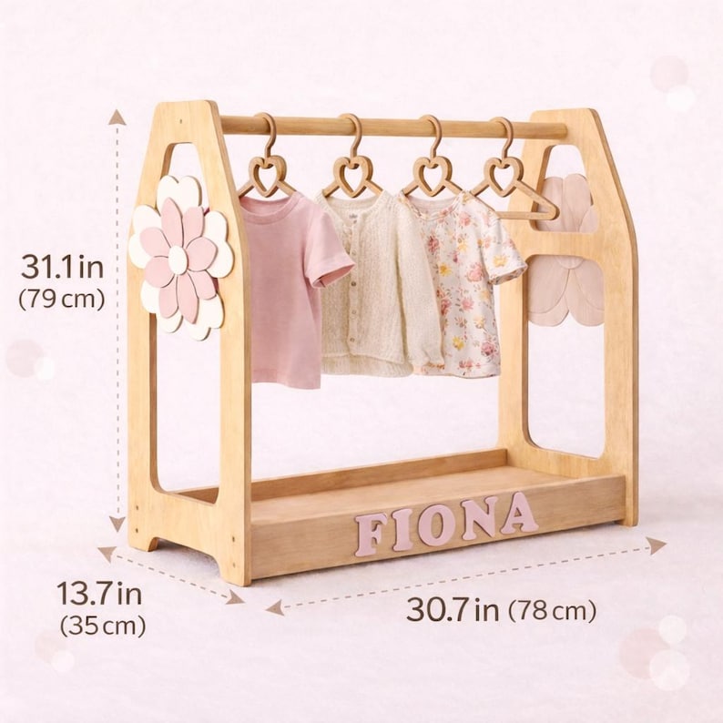 May include: A wooden children's clothing rack, 30.7 inches (78 cm) wide and 31.1 inches (79 cm) tall, with a flower accent and the name "FIONA" in pink letters. It holds several small garments on heart-shaped hangers.