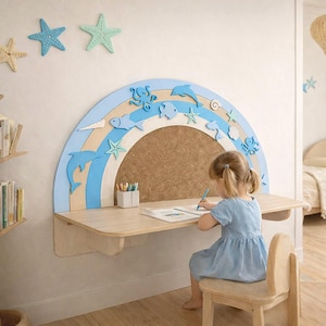 May include: A light wood desk with a blue and white rainbow-shaped bulletin board. The board is decorated with sea creatures. Above the desk are starfish wall decorations. A young child is sitting at the desk, drawing with a blue pen.