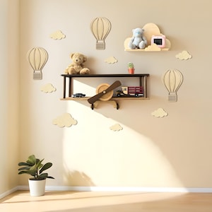Peut inclure: Décoration murale de chambre d'enfant avec une étagère en forme d'avion, des découpes de montgolfières et des nuages. Un ours en peluche, un jouet bleu et un petit appareil photo rose sont sur les étagères. Une plante en pot est au sol.
