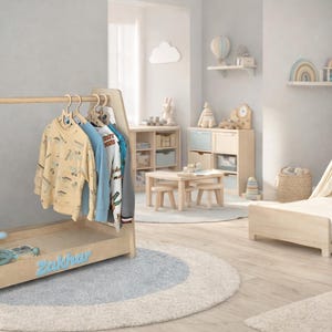 May include: A light wood children's room with a clothes rack, teepee, and bed. The clothes rack has a rocket ship design and the name "Zakhar" in blue. Several shirts hang on the rack. The room has a light blue and white color scheme.