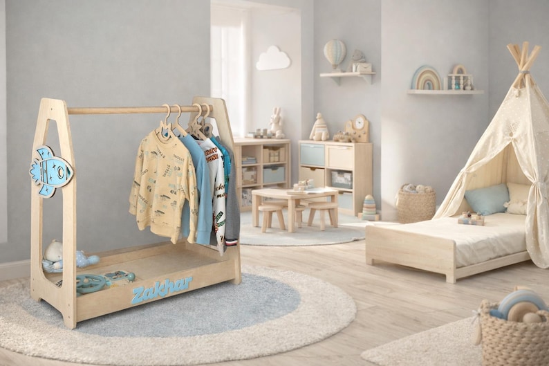 May include: A light wood children's room with a clothes rack, teepee, and bed. The clothes rack has a rocket ship design and the name "Zakhar" in blue. Several shirts hang on the rack. The room has a light blue and white color scheme.