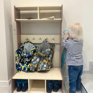 May include: A light-colored wooden hall tree featuring an upper shelf with two compartments, a central panel with five silver metal hooks for hanging items, and a lower bench section with three open cubbies for storing footwear.