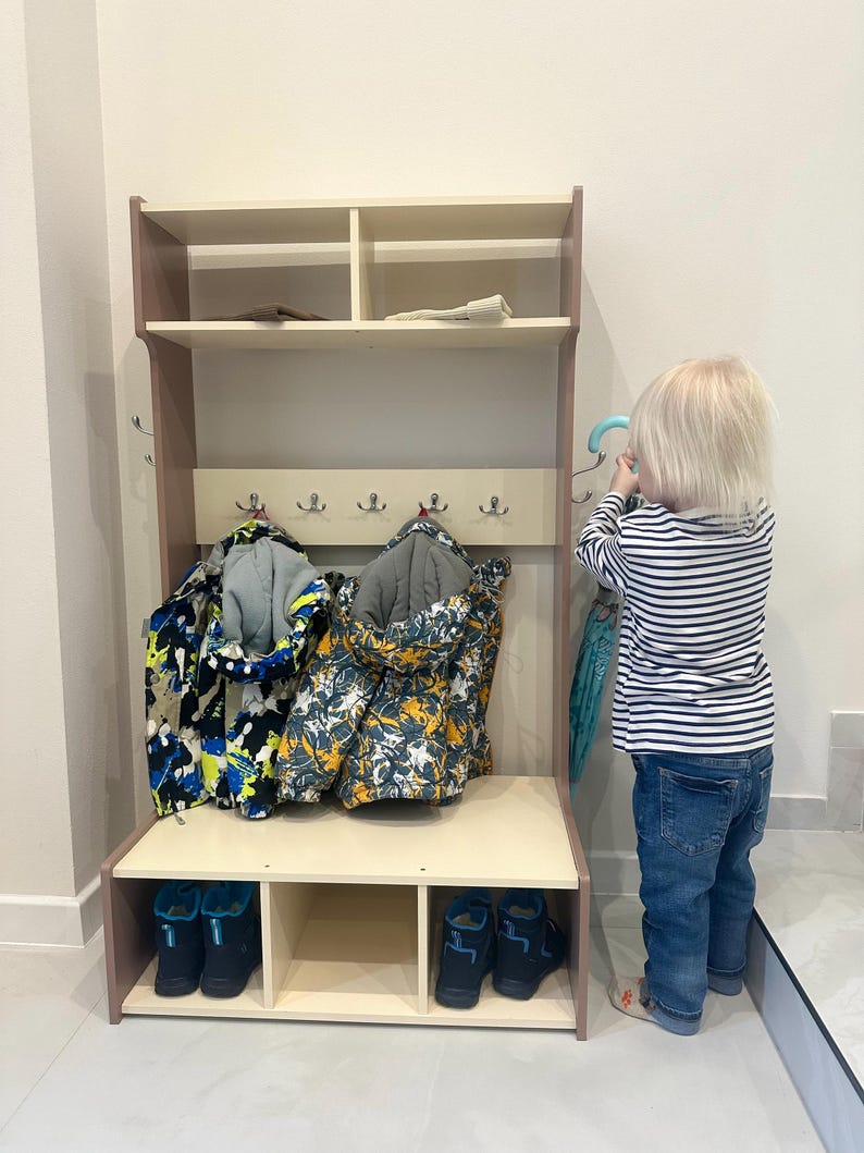 May include: A light-colored wooden hall tree featuring an upper shelf with two compartments, a central panel with five silver metal hooks for hanging items, and a lower bench section with three open cubbies for storing footwear.