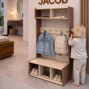 May include: A wooden entryway storage unit with a top shelf, a middle section featuring four metal hooks, and a bottom bench with three open cubbies for shoes. The name JACOB is mounted in large brown letters on the wall above the storage unit.