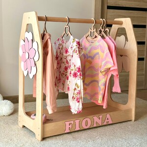 May include: Wooden children's clothing rack with a flower and heart-shaped cutouts. The rack holds several garments, including a pink long-sleeve shirt, a floral dress, and a colorful striped t-shirt. The name "FIONA" is spelled out in pink letters.
