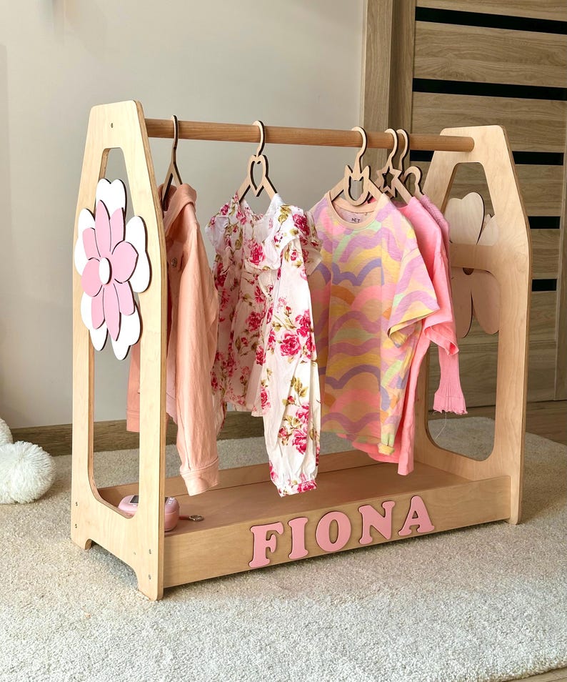 May include: Wooden children's clothing rack with a flower and heart-shaped cutouts. The rack holds several garments, including a pink long-sleeve shirt, a floral dress, and a colorful striped t-shirt. The name "FIONA" is spelled out in pink letters.
