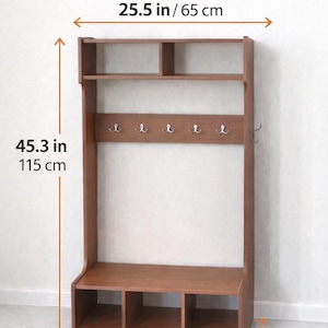 May include: A brown wooden hall tree with a top shelf, five silver hooks, and a bottom bench with three storage compartments. The unit measures 25.5 inches wide, 45.3 inches high, and 15.7 inches deep, as indicated by the text Dimensions.