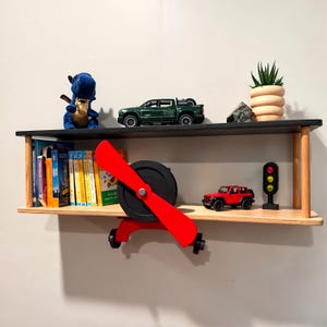 May include: A wooden airplane shelf with two shelves. The top shelf has a green toy truck, a blue stuffed animal, and a small potted plant. The bottom shelf has a red toy car, a traffic light, and a collection of colorful books.