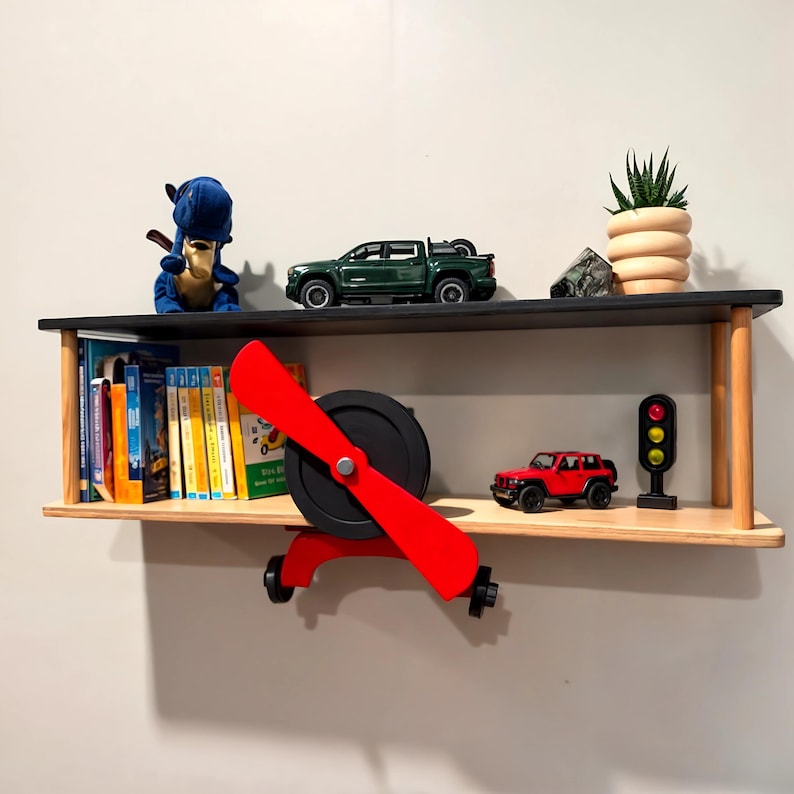 May include: A wooden airplane shelf with two shelves. The top shelf has a green toy truck, a blue stuffed animal, and a small potted plant. The bottom shelf has a red toy car, a traffic light, and a collection of colorful books.