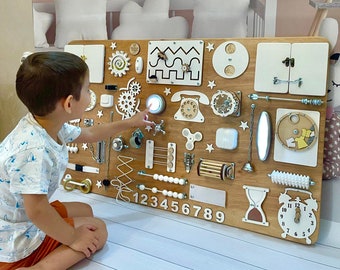 May include: A wooden activity board with various textures and activities for children. It features a light switch, a phone, a zipper, a clock, a bead maze, a spinning wheel, and a number line. The board is painted in natural wood tones and white.