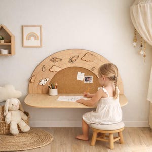 Puede incluir: Un escritorio de madera en forma de arcoíris con recortes de cohetes y nubes, un corcho y un taburete pequeño. Una niña con un vestido blanco dibuja. Un cuadro de arcoíris enmarcado cuelga de la pared.