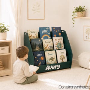 May include: A dark green wooden floor-standing book display shelf featuring three slanted tiers for book storage. The front panel is personalised with the white text Avery. The shelf holds nine children's books with various educational titles.