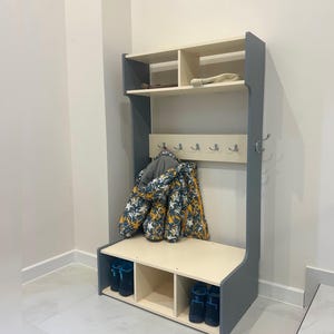 May include: A gray and cream hall tree featuring an upper shelf, a central panel with five silver hooks, and a lower bench with three shoe compartments. Text reads: By default, the wardrobe comes with three shoe compartments. If you would prefer two instead, just let us know - we'll be happy to customize it for you.