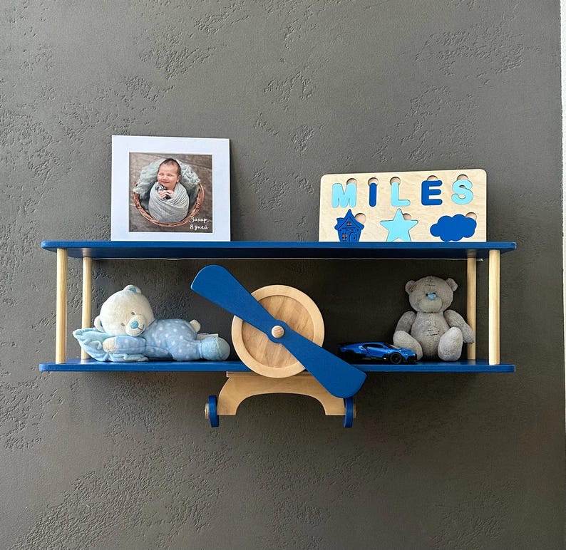 May include: Blue airplane-shaped shelf with two shelves. The top shelf has a wooden name puzzle that says "MILES" and a few wooden shapes. The bottom shelf has a teddy bear, a toy car, and a framed photo of a baby.