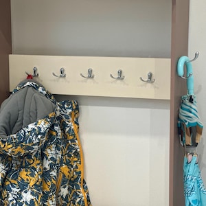 May include: A wall-mounted coat rack featuring a light beige horizontal panel with five silver-toned double hooks. A patterned jacket with blue, yellow, and white abstract shapes hangs on the left, while a light blue umbrella hangs on the right.