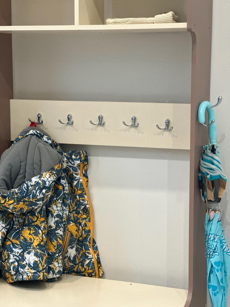 May include: A wall-mounted coat rack featuring a light beige horizontal panel with five silver-toned double hooks. A patterned jacket with blue, yellow, and white abstract shapes hangs on the left, while a light blue umbrella hangs on the right.