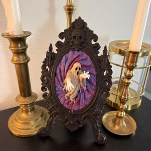 May include: A dark brown, ornate, oval-shaped frame featuring a skull at the top. Inside the frame, a three-dimensional, off-white ghost figure with outstretched arms emerges from a swirling, textured background of deep purple and red hues.