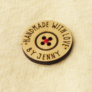 Puede incluir: Una etiqueta redonda de madera con el texto "Handmade with love. By Jenny" y un botón rojo en el centro.