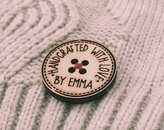May include: A wooden button with the text "Handcrafted with love by Emma" in black lettering. The button has a red thread sewn through four holes in the center.