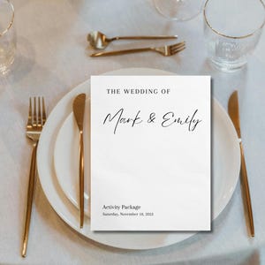 Może przedstawiać: Biała prostokątna karta z planem atrakcji weselnych z czarnym napisem THE WEDDING OF Mark & Emily, Activity Package, Saturday, November 18, 2023, umieszczona na białym talerzu obiadowym ze złotymi sztućcami i przezroczystymi szklankami.