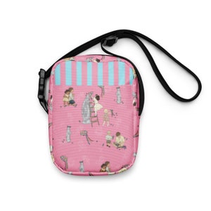 Puede incluir: Bolso bandolera rosa con correa y hebilla negras. El bolso presenta un patrón repetido de niños, gatos y ramos de flores. El bolsillo superior tiene un diseño de rayas azules y blancas.