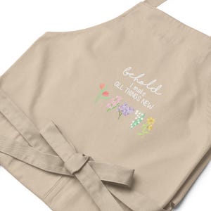 May include: Beige kitchen apron with a white embroidered quote "behold I make all things new" and a row of colorful flowers.