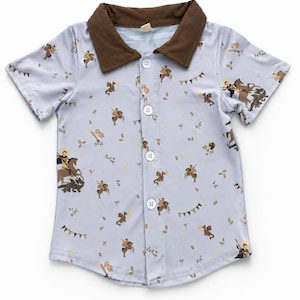 May include: A light blue short-sleeve button-down shirt featuring a repeating pattern of knights on horseback, dragons, swords, and pennant banners. The shirt has a solid brown collar and four white buttons down the center front placket.